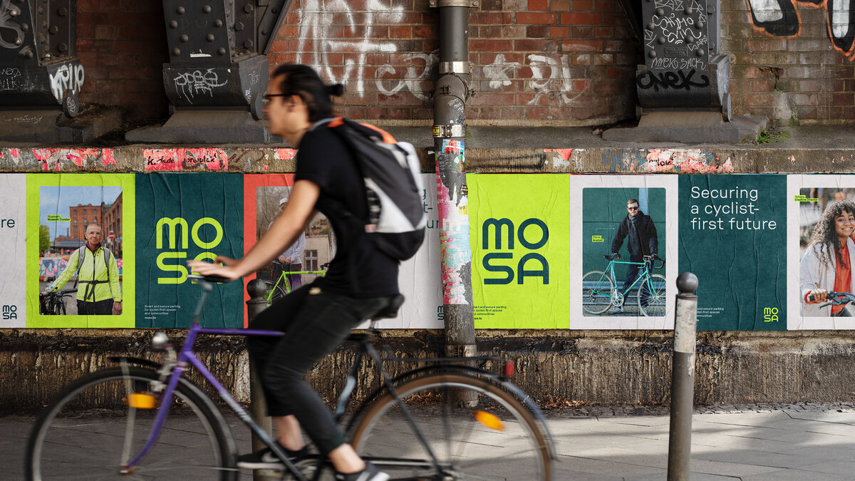 Photograph of young man with backpack and a ponytail cycling past Mosa out of home advertising.